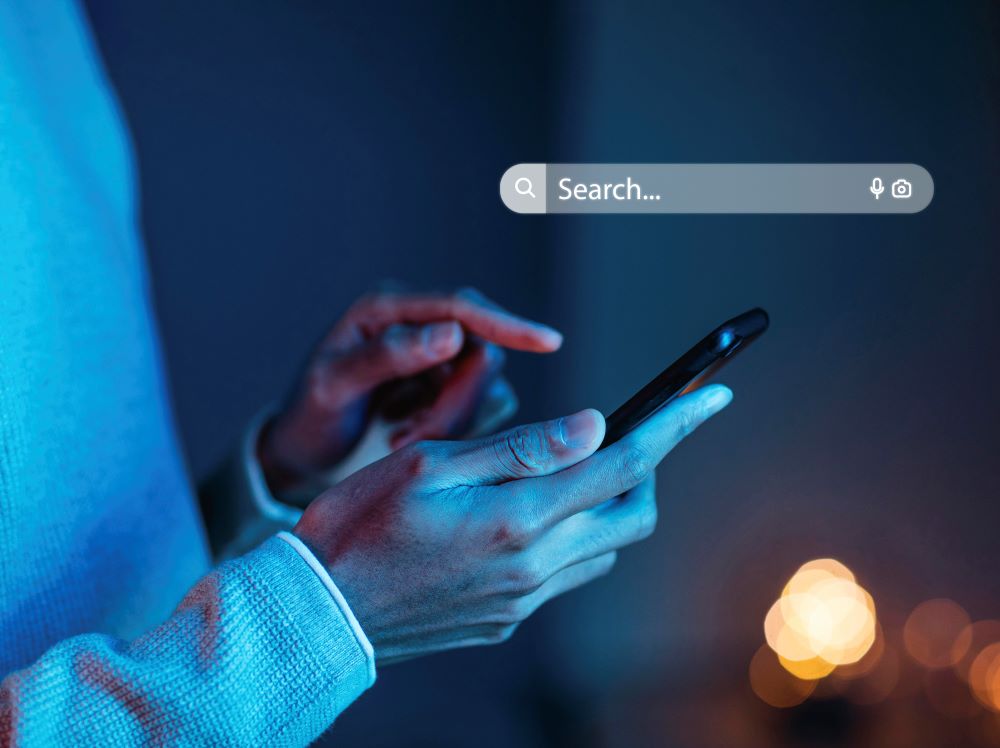 A imagem mostra uma pessoa segurando um smartphone com uma das mãos e tocando a tela com a outra. A cena está iluminada por uma luz azul suave, sugerindo um ambiente noturno ou com pouca iluminação. No topo da imagem, há uma barra de busca flutuante com o ícone de uma lupa e a palavra "Search..." indicando que a pessoa está prestes a realizar uma pesquisa na internet, realçando a importância do SEO.