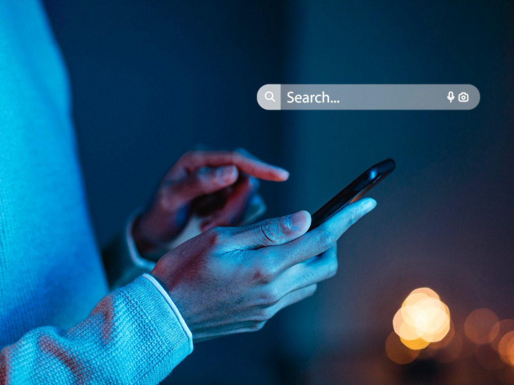 Um close-up de uma pessoa segurando um smartphone com ambas as mãos, concentrada na tela. A pessoa está usando um suéter de manga comprida. No canto superior da imagem, há uma barra de pesquisa translúcida com a palavra "Search..." e ícones de microfone e câmera. A iluminação é azulada, com luzes desfocadas ao fundo, criando um ambiente moderno e tecnológico.