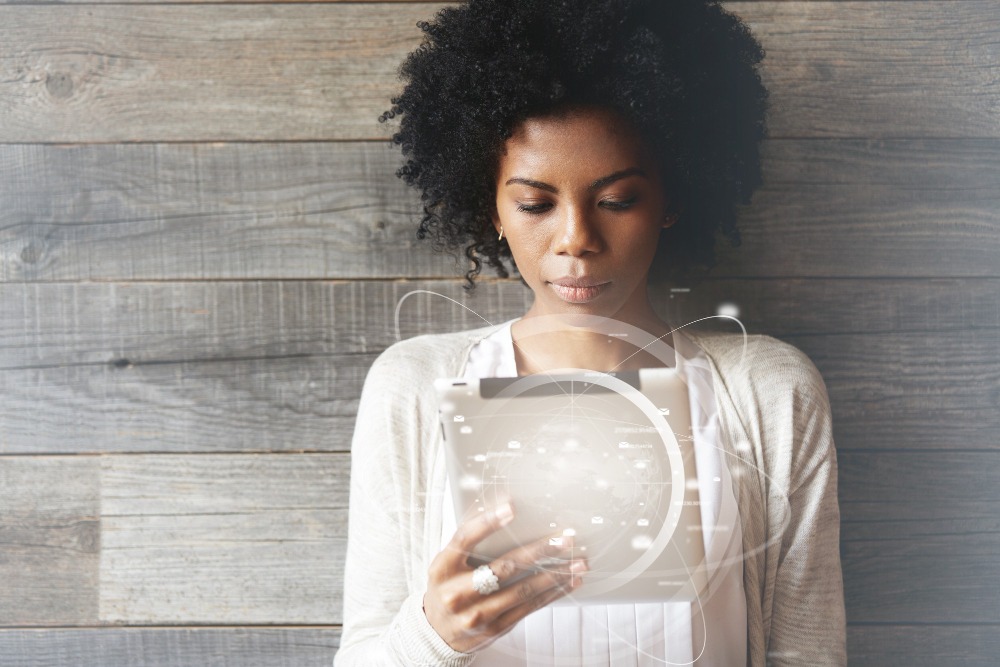 Uma mulher de pele escura e cabelo afro está concentrada olhando para um tablet, que ela segura com ambas as mãos. Ela veste uma camiseta branca e um cardigã cinza claro. O fundo é uma parede de madeira cinza, e há um efeito digital sobre a imagem, com gráficos holográficos de dados e informações flutuando ao redor do tablet, sugerindo que ela está analisando ou interagindo com informações digitais avançadas. A expressão dela é séria e focada, destacando a concentração no que está fazendo.