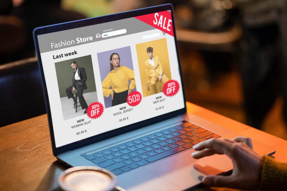 Um close de um laptop aberto mostra uma loja online de moda com uma seção de promoções. Três produtos estão em destaque: um terno feminino preto com 80% de desconto, um suéter de lã amarelo com 50% de desconto, e um terno masculino amarelo com 80% de desconto. Uma mão está navegando na página, e o laptop está em uma mesa de madeira com uma xícara de café ao lado. O fundo está desfocado, destacando a atenção na tela.