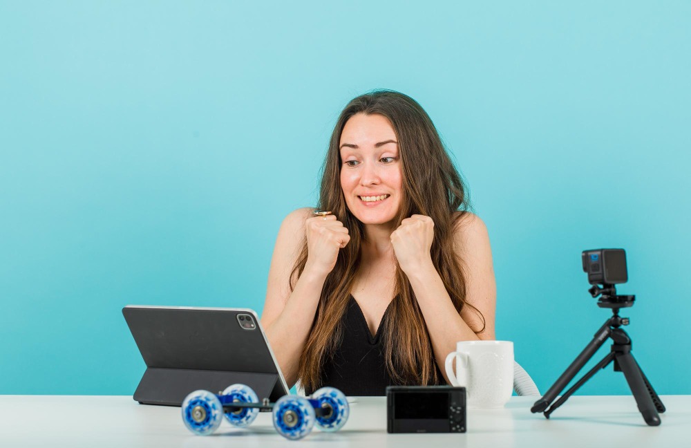 Uma mulher com expressão de felicidade e entusiasmo sentada à mesa com equipamentos de gravação de vídeo, incluindo uma câmera, um tripé, um tablet e um par de patins. O fundo é azul claro.