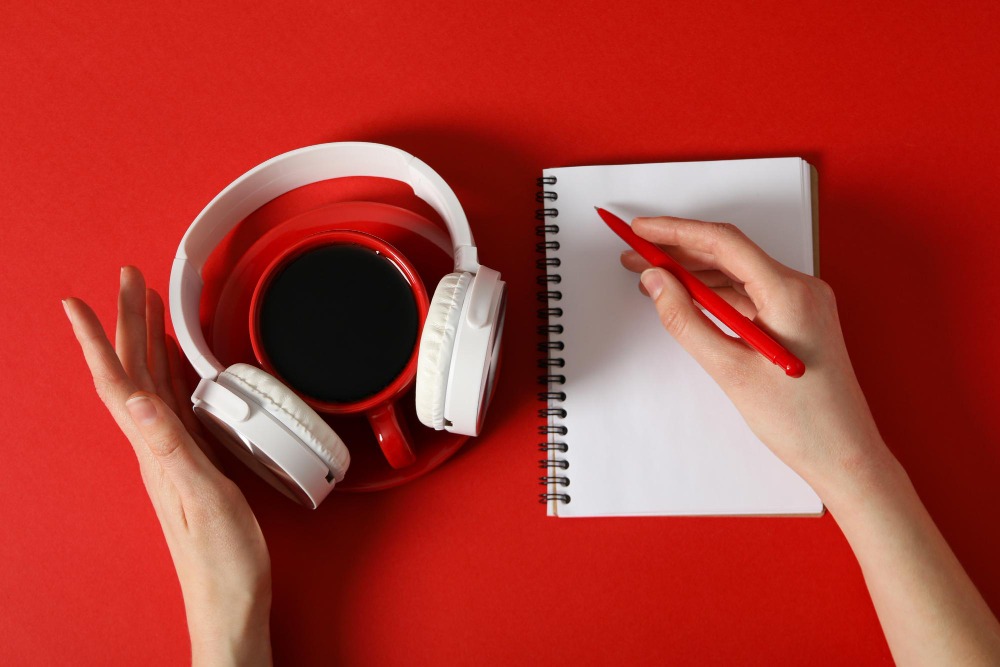 Uma mesa vermelha com uma xícara de café preto, fones de ouvido brancos ao redor da xícara, e uma mão segurando um caderno aberto com uma caneta vermelha pronta para escrever. Esta imagem sugere preparação para estudar ou tomar notas sobre "Quantos é pronome".