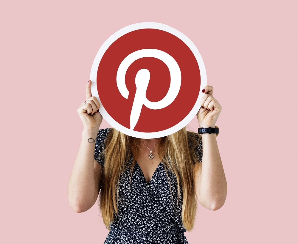 Uma mulher loira segurando um grande logo redondo do Pinterest na frente do rosto. O fundo é rosa claro. Pinterest fotos.