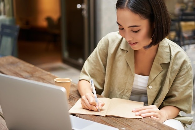 Portrait of young asian woman working on laptop making notes writing down while attending online lesson work meeting